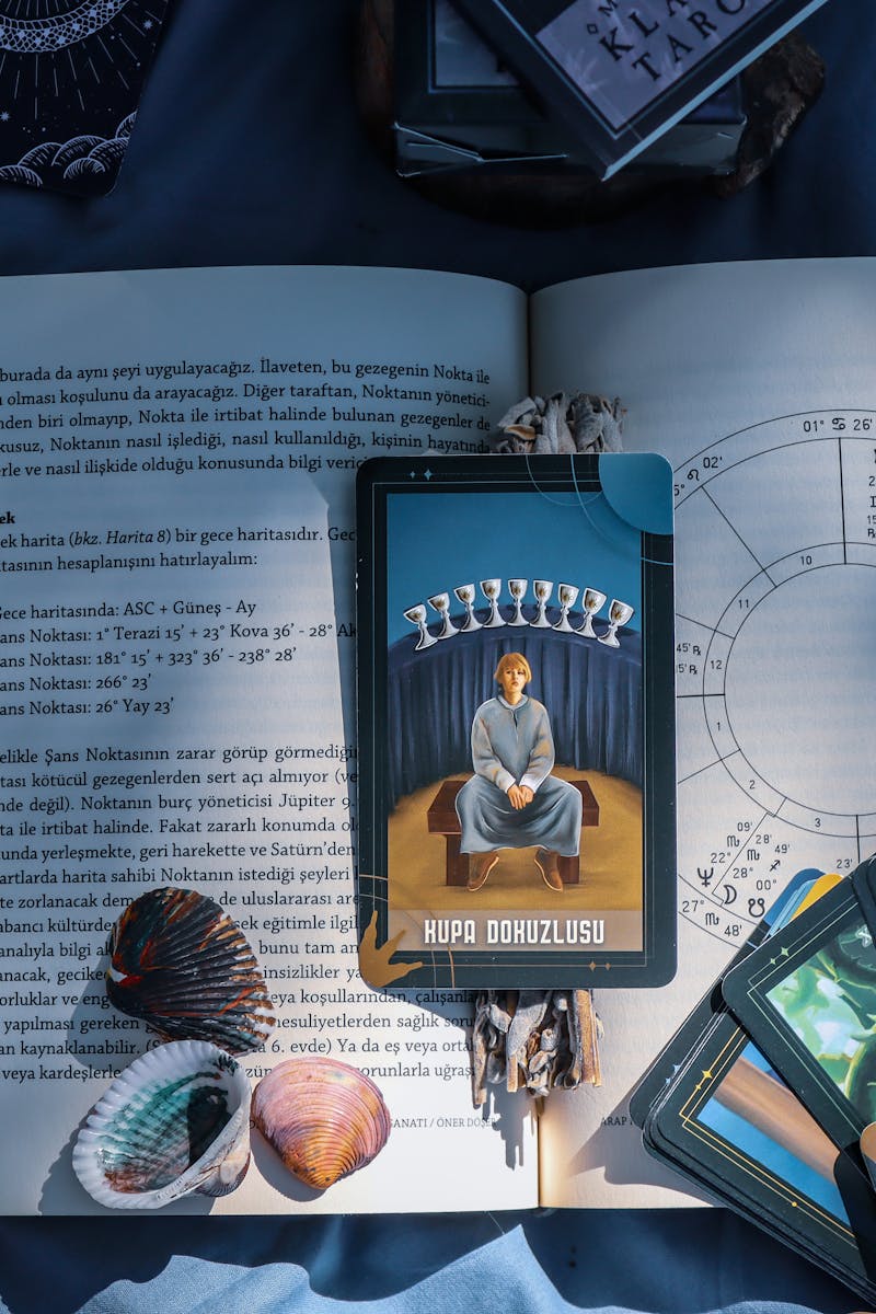 An artistic flat lay of tarot cards and astrology on a book with seashells, featuring Kupa Dokuzlusu card.